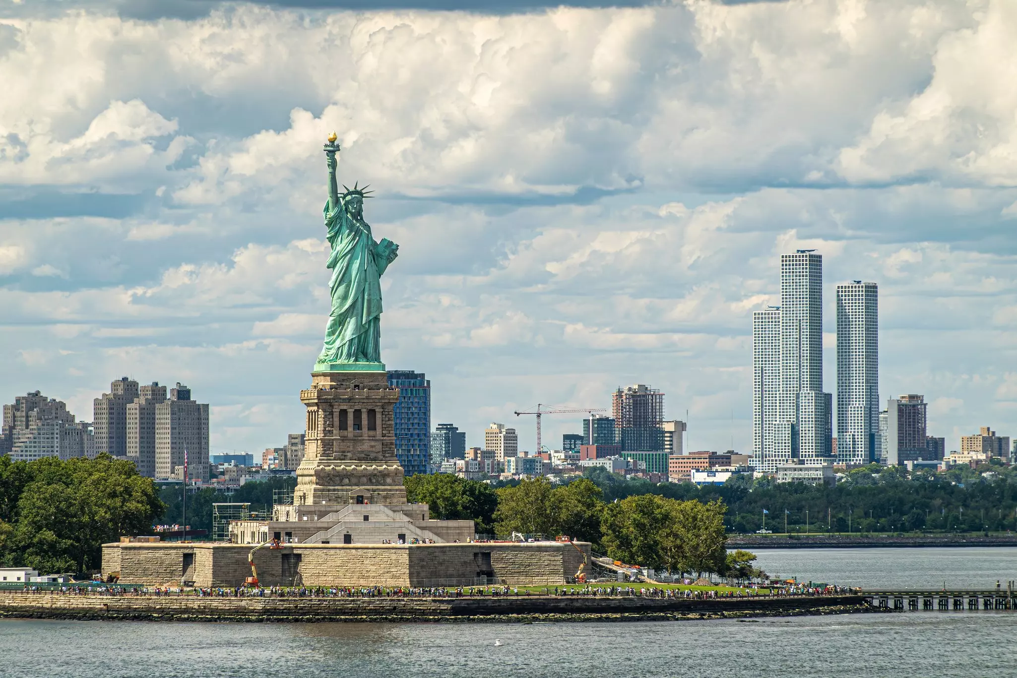 A monumental statue of a woman, turned green through oxidation, is placed on a plinth on an island in the harbor of a city. Towers of the city are visible on the shore.