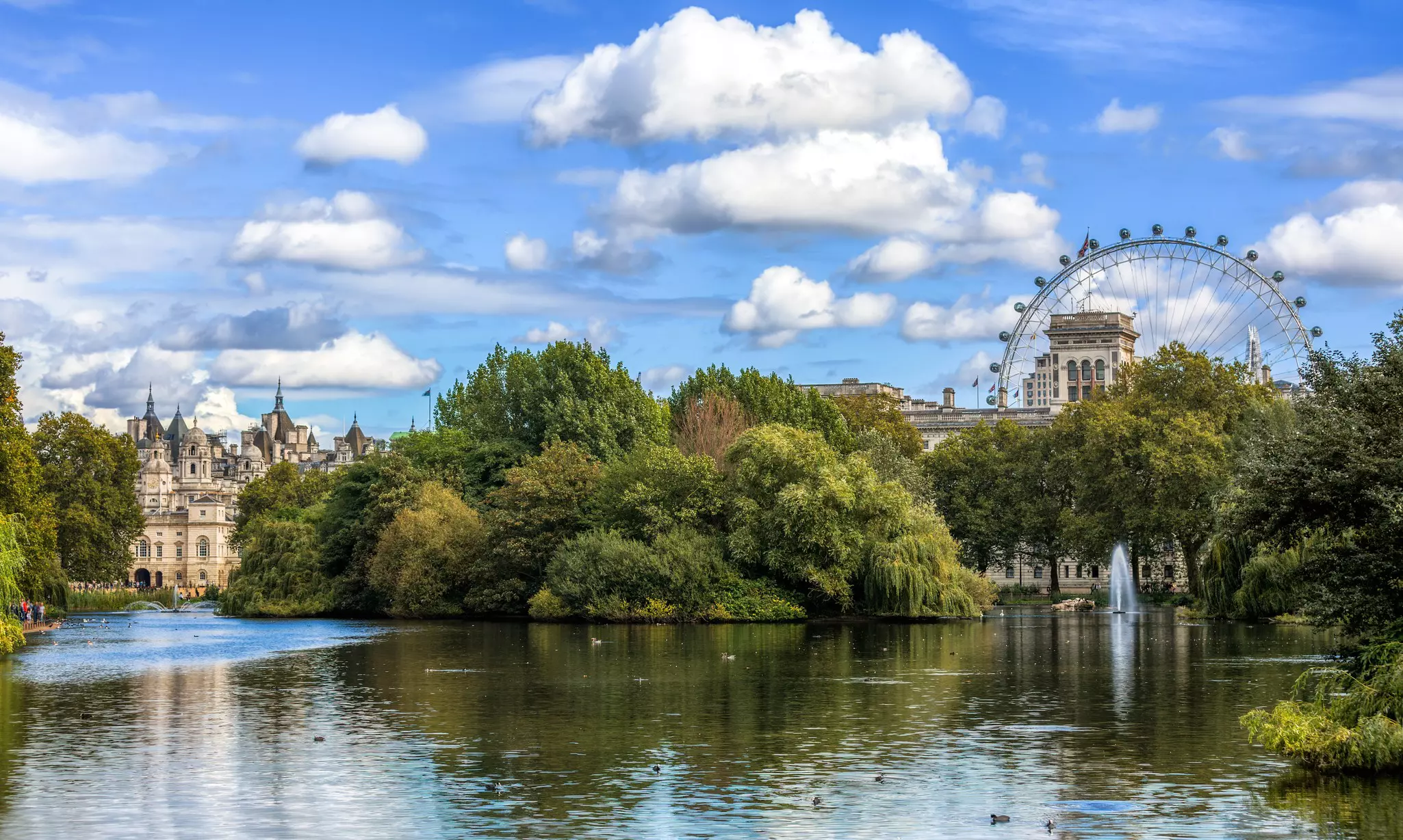 Pond and trees in a city park with ferris wheel and historic ornate buildings in the background on a partly sunny day.