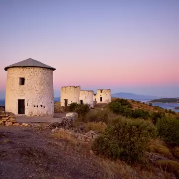 Four round white stone mill towers, one with a conical roof, on a scrubby ridge over a bay with boats under a pink sky