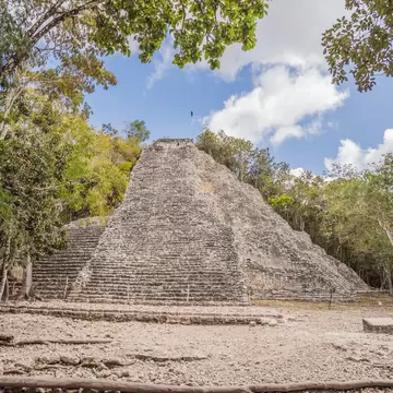 Steep stepped stone pyramid with a small temple on top, framed by trees under a blue sky with clouds