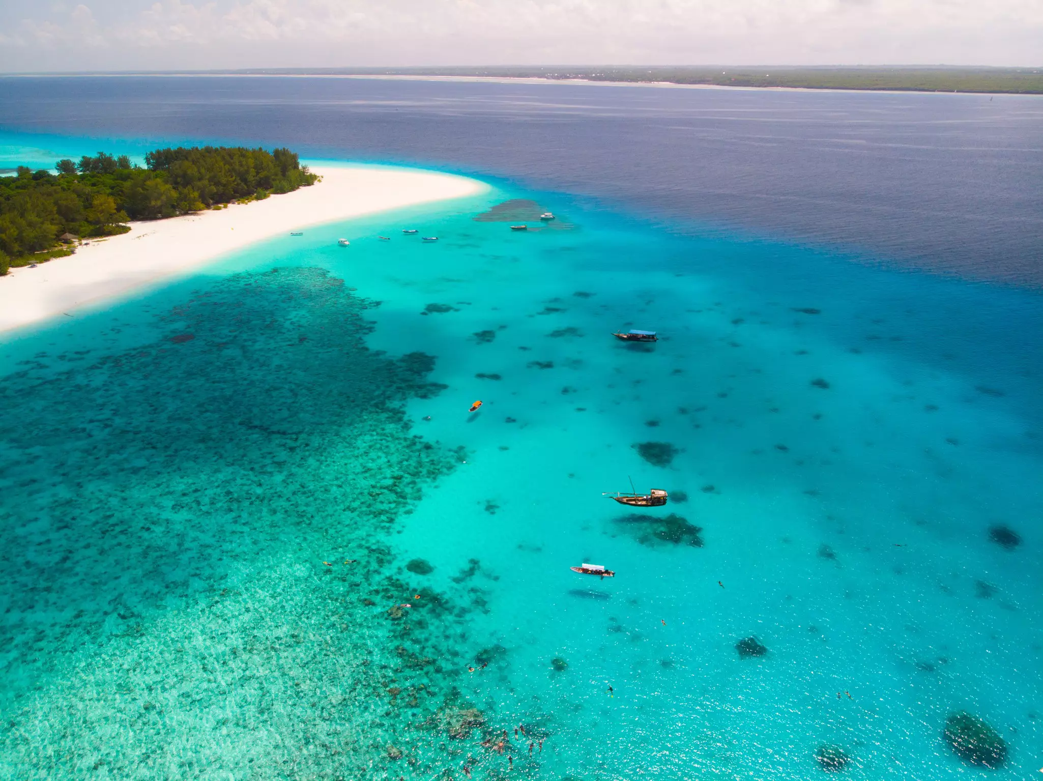 Aerial view of clear, bright blue ocean with a few boats and narrow peninsula of white sand beach and forest to the left on a sunny day.