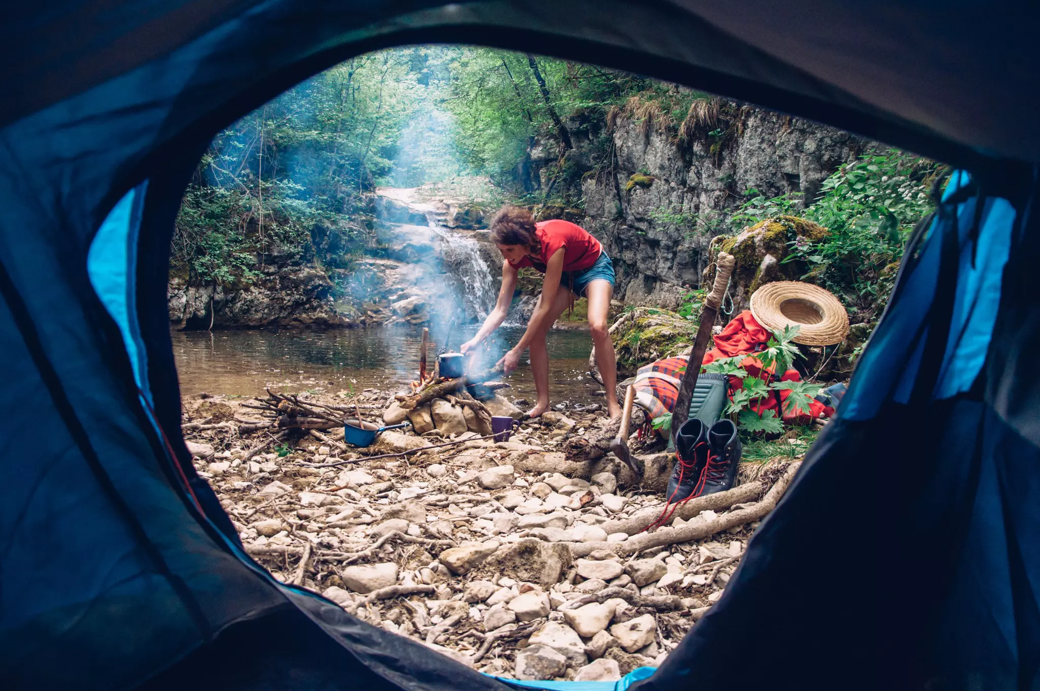 A woman prepares a campfire, viewed through the opening of the tent. She has walking boots and cookery gear nearby