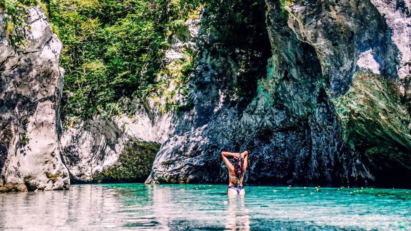 A woman slicks back her wet hair after swimming in the turquoise Soča River with hulking rocks in front of her