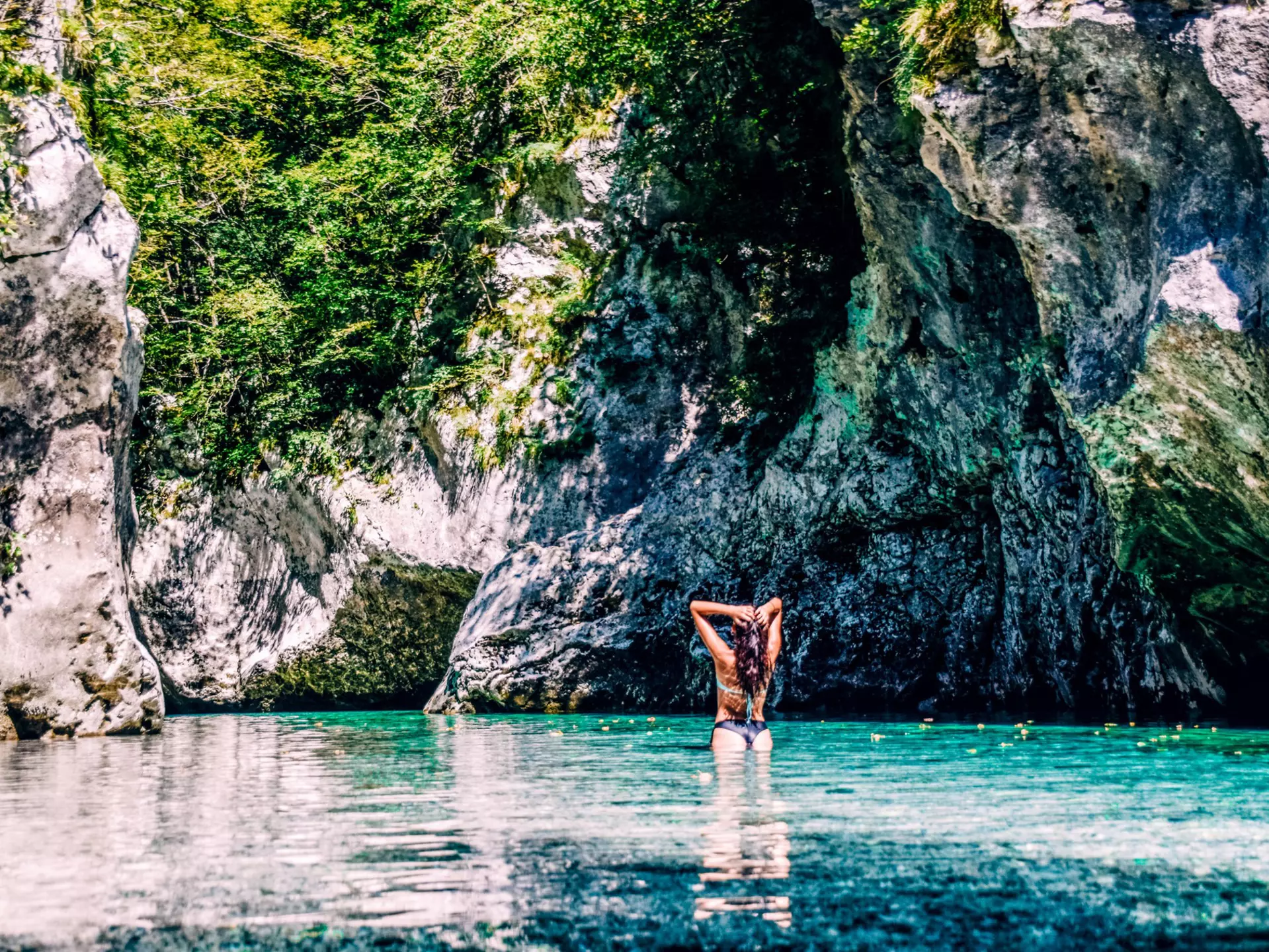A woman slicks back her wet hair after swimming in the turquoise Soča River with hulking rocks in front of her