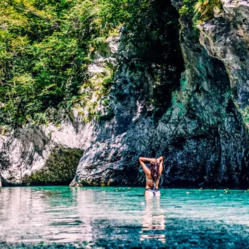 A woman slicks back her wet hair after swimming in the turquoise Soča River with hulking rocks in front of her