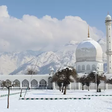 The white-domed Hazratbal mosque and its minaret in Srinagar, Kashmir, with snow-covered grounds, trees and mountains.