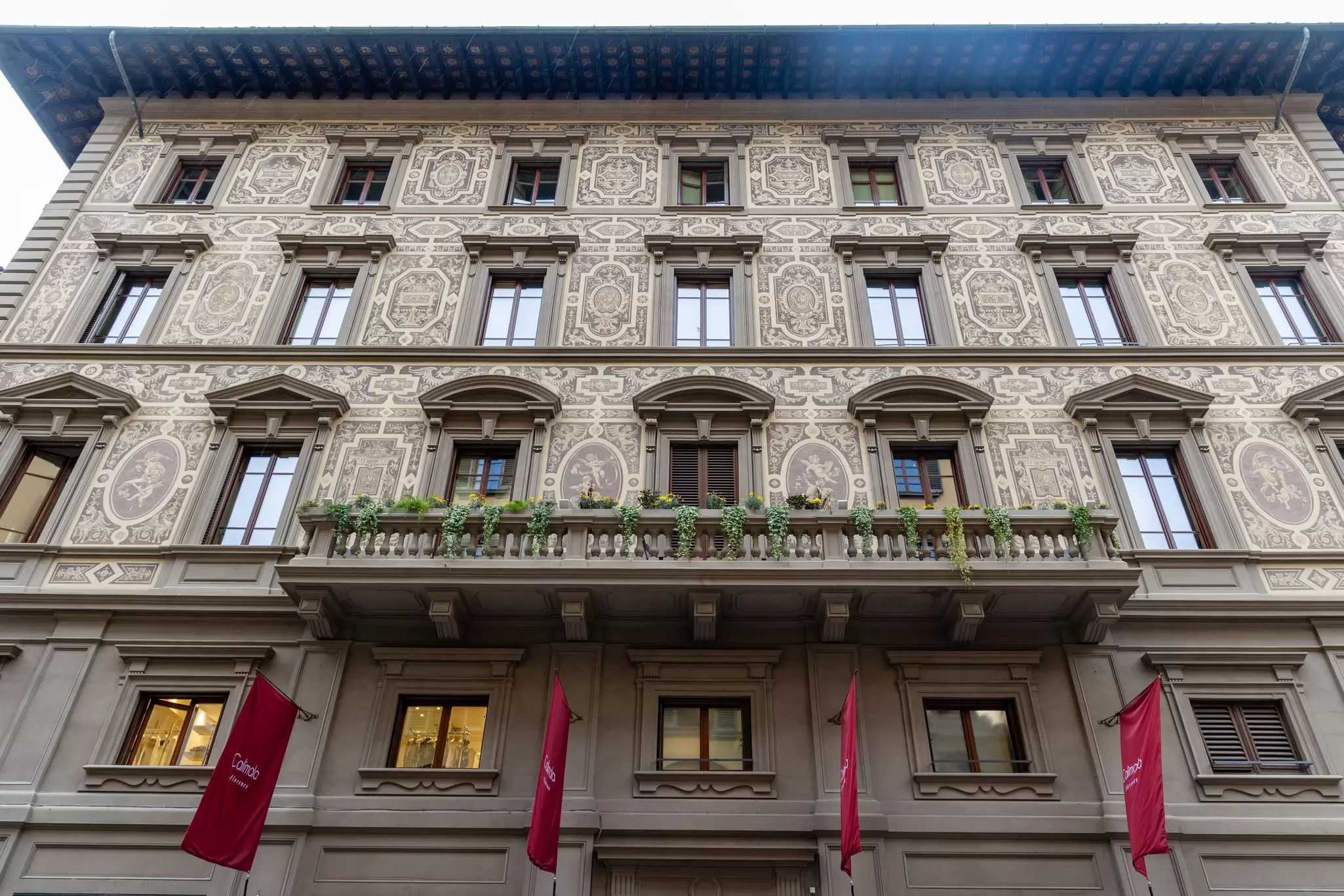 Facade of a grand renaissance-style building with intricate patterns, a balcony, and red flags hanging over the doors: Hotel Calimala, Florence