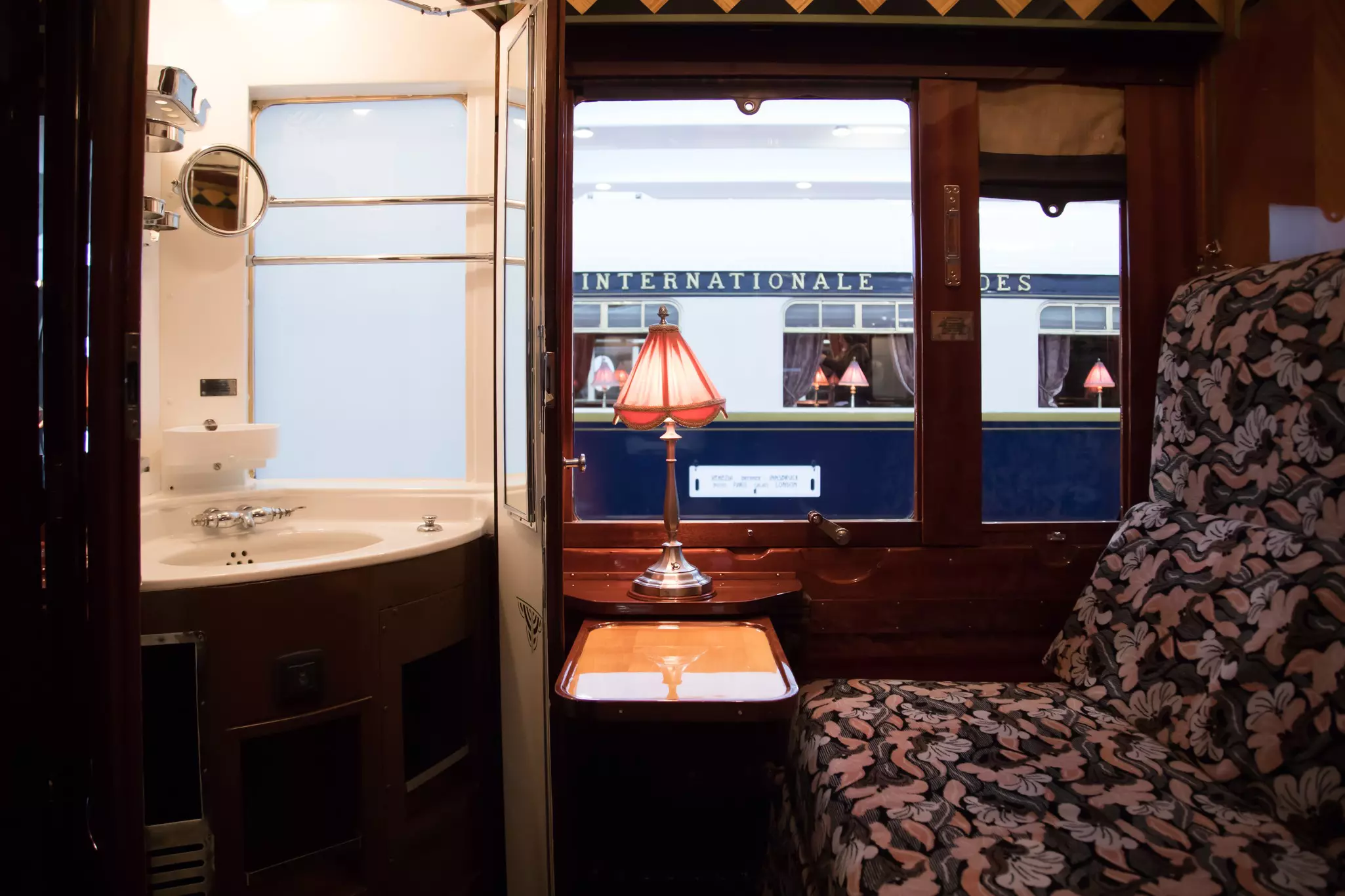 A luxurious train interior, with patterned seats, a lamp on a table and a small washroom.