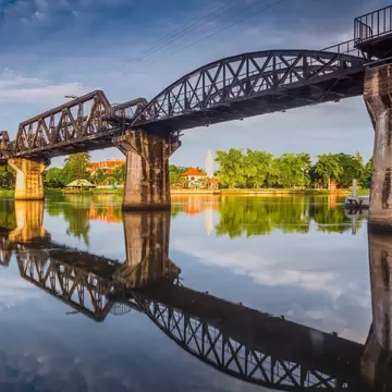 Steel truss railway bridge on concrete piers crossing a calm river in Kanchanaburi, mirrored in the water.