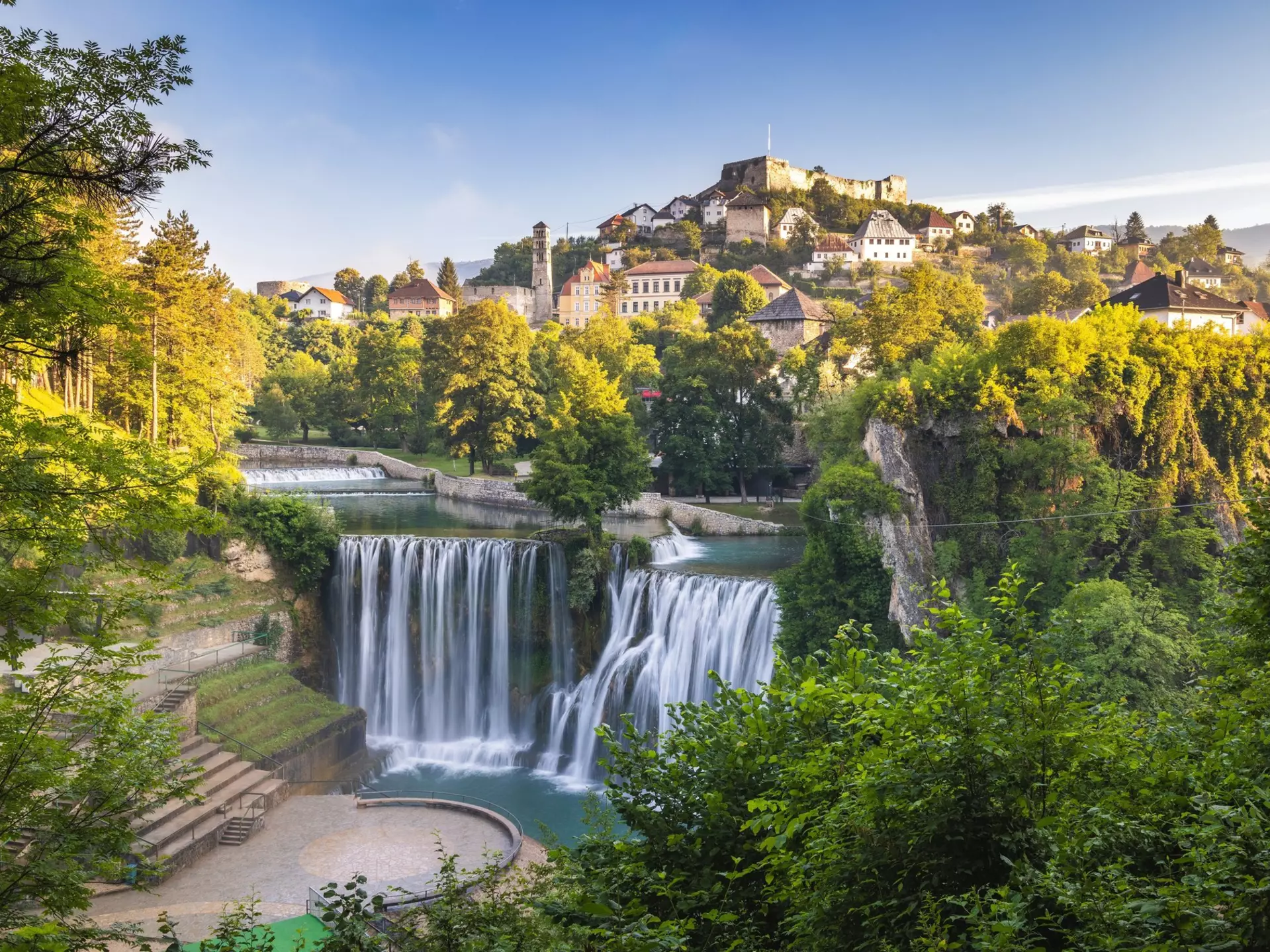 A waterfall cascades into a pool below a hilltop town that glows in the spring sunshine.