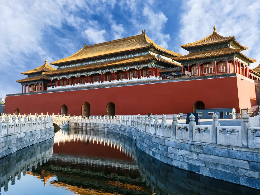 A red building with a peaked roof is on one side of a stone wall lining a waterway.