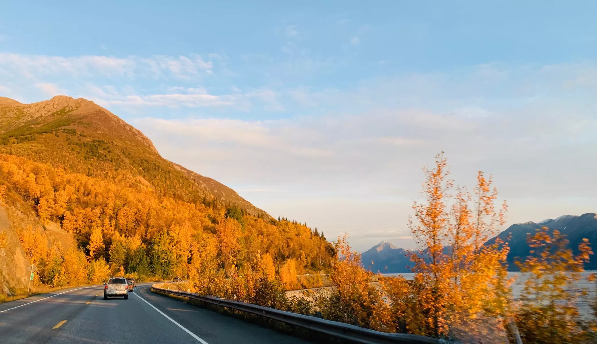 A car drives on a highway surrounded by trees in fall foliage. An inlet is one side of the highway.