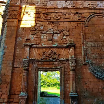 An ornately carved red sandstone doorway in a ruined mission wall, a frieze of small figures above it and green parkland visible through the opening.