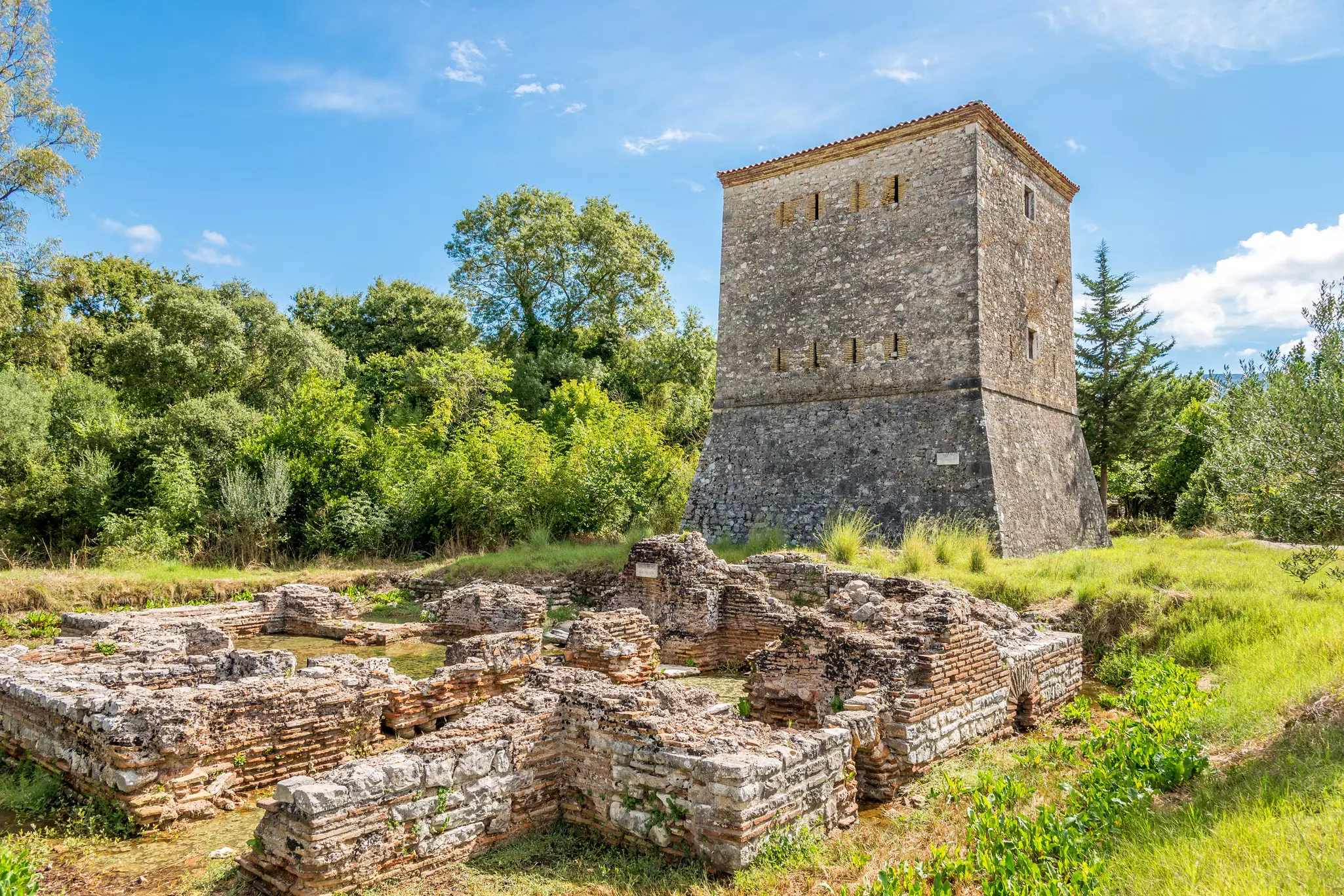 A Venetian tower in Butrint, with grass, trees and a mostly blue sky