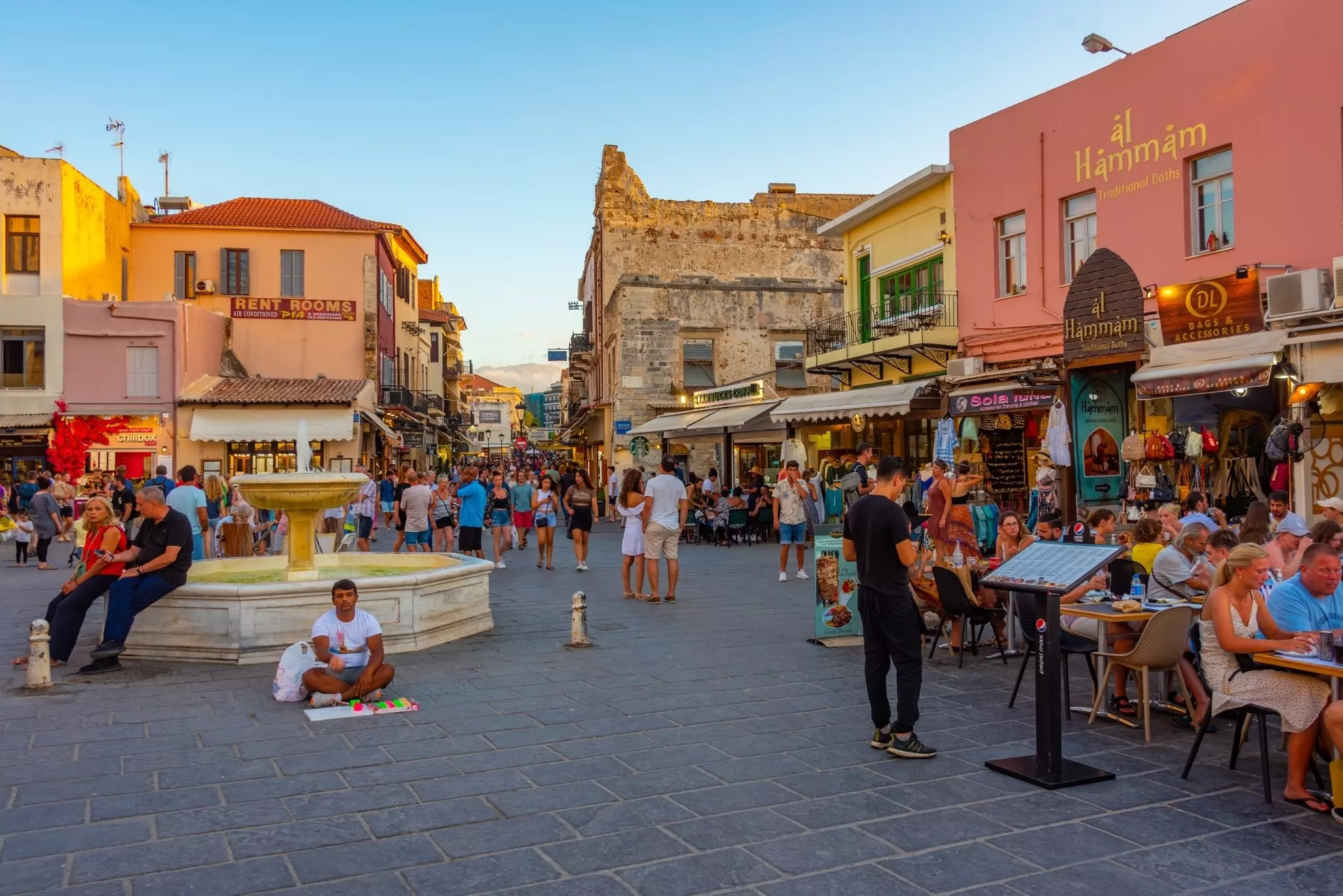 People at outdoor cafes and walking through an open plaza with a white marble fountain.