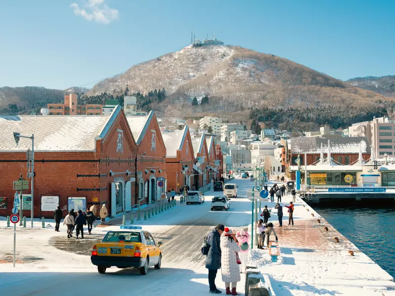 A view of people walking by brick waterhouses at a port in a city. Snow covers the ground, and mountains rise in the distance.