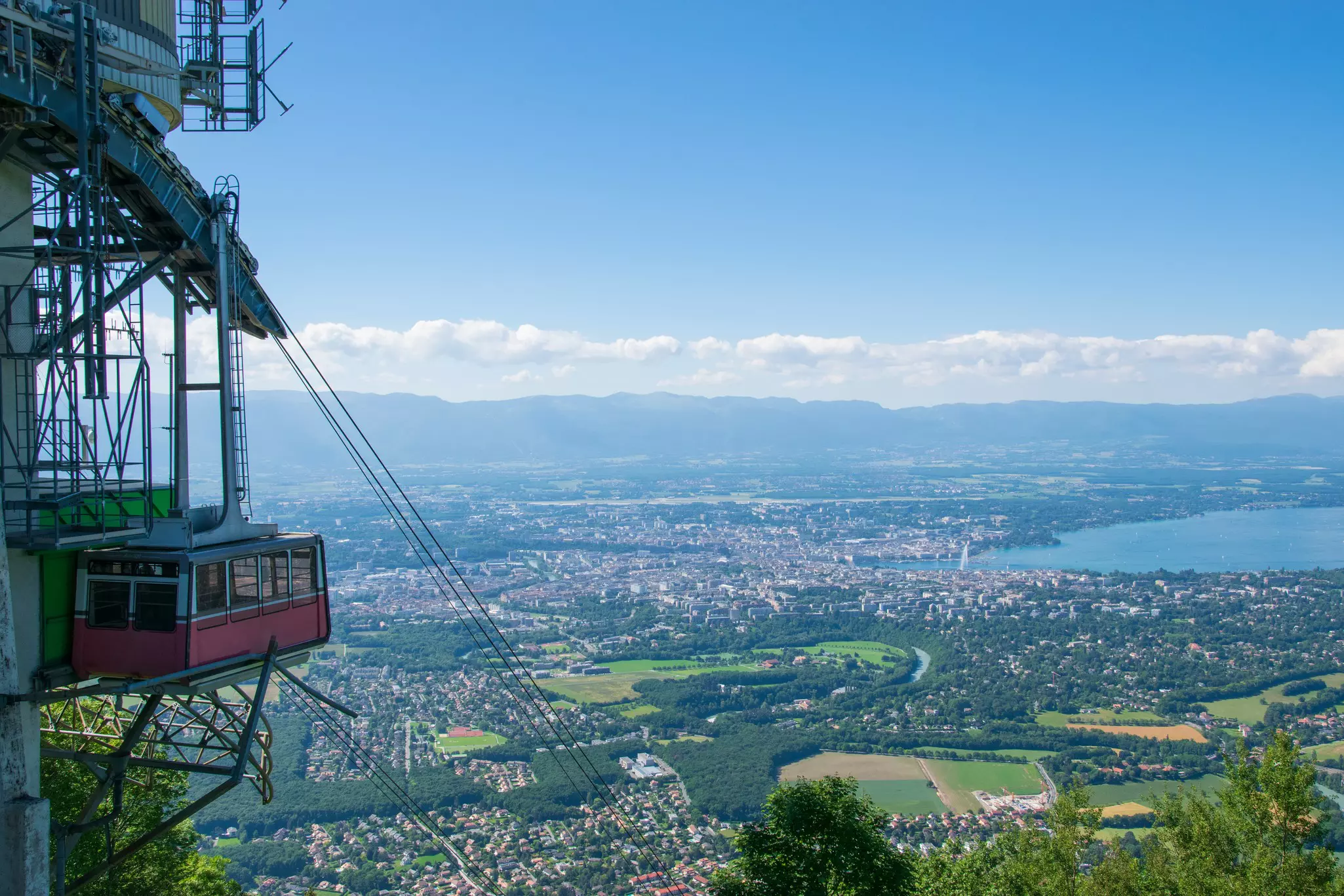 A cable car gondola is docked at the summit of a mountain. Dramatic views of a city in a green valley are seen in the distance.