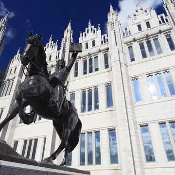 A bronze equestrian statue with the rider holding a charter aloft, in front of a white granite Gothic facade of pinnacles, spires and tall mullioned windows.
