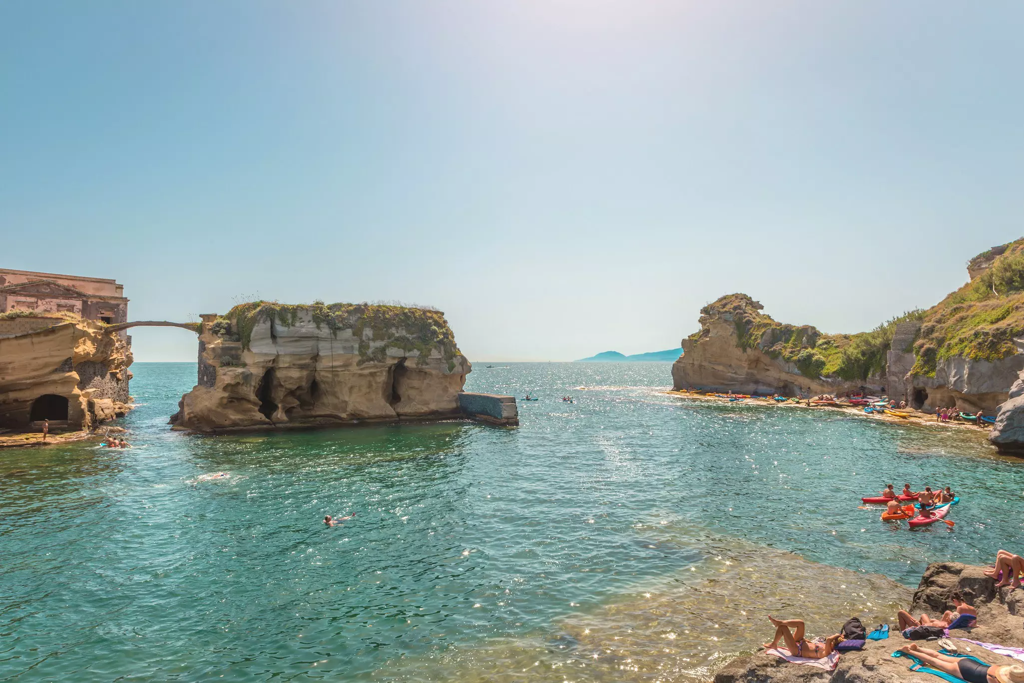 A wide view of a shallow cove, with rock islets in the water, and kayakers seen by the shore to the right.