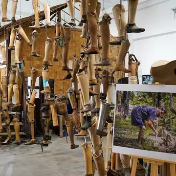 Dozens of hanging prosthetic legs fill an exhibition room, with a photo of a man digging on an easel