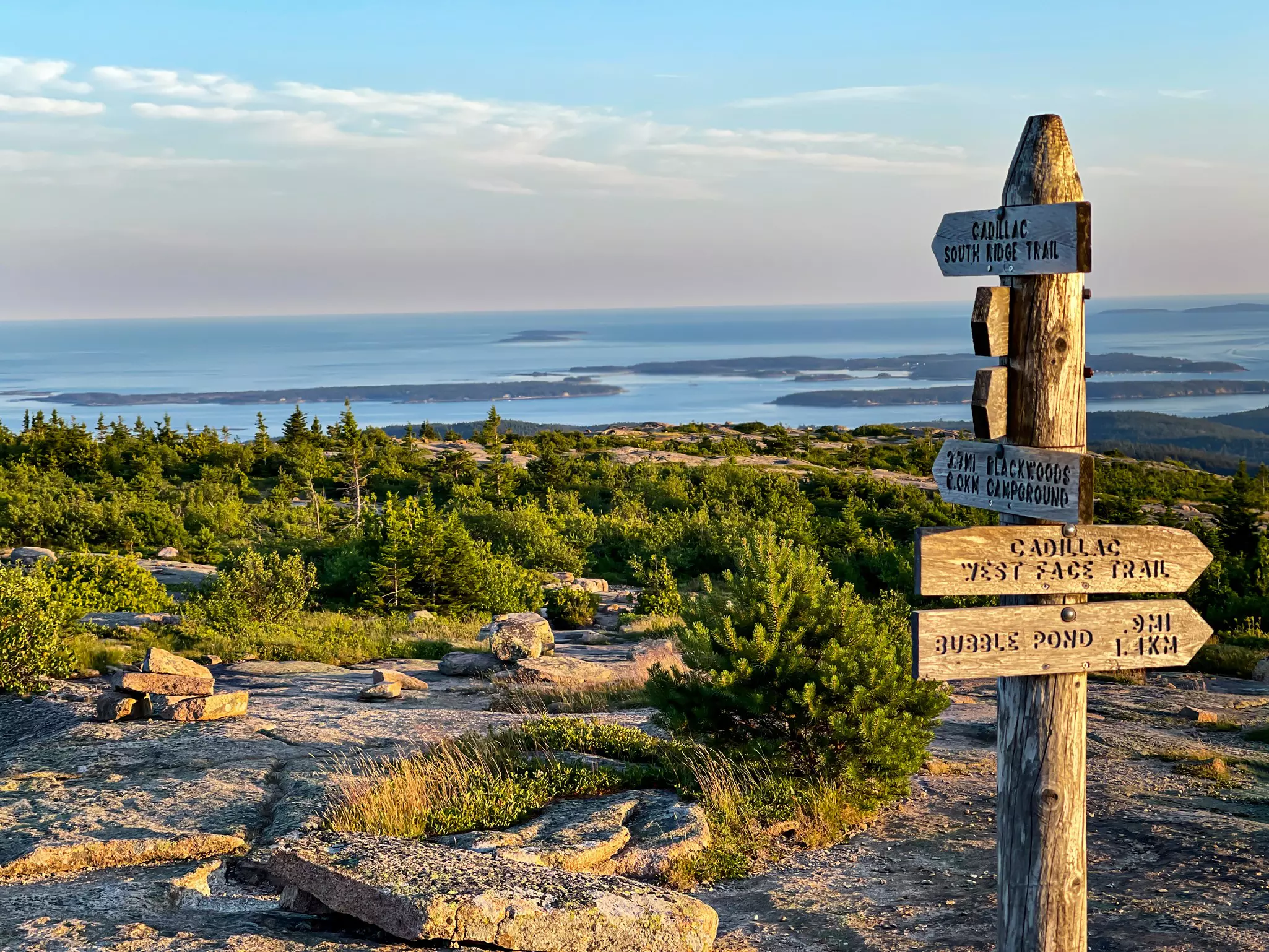 A rocky summit of a mountain with a wooden sign showing the various trail routes. Views stretch out over the ocean and islands in the distance.