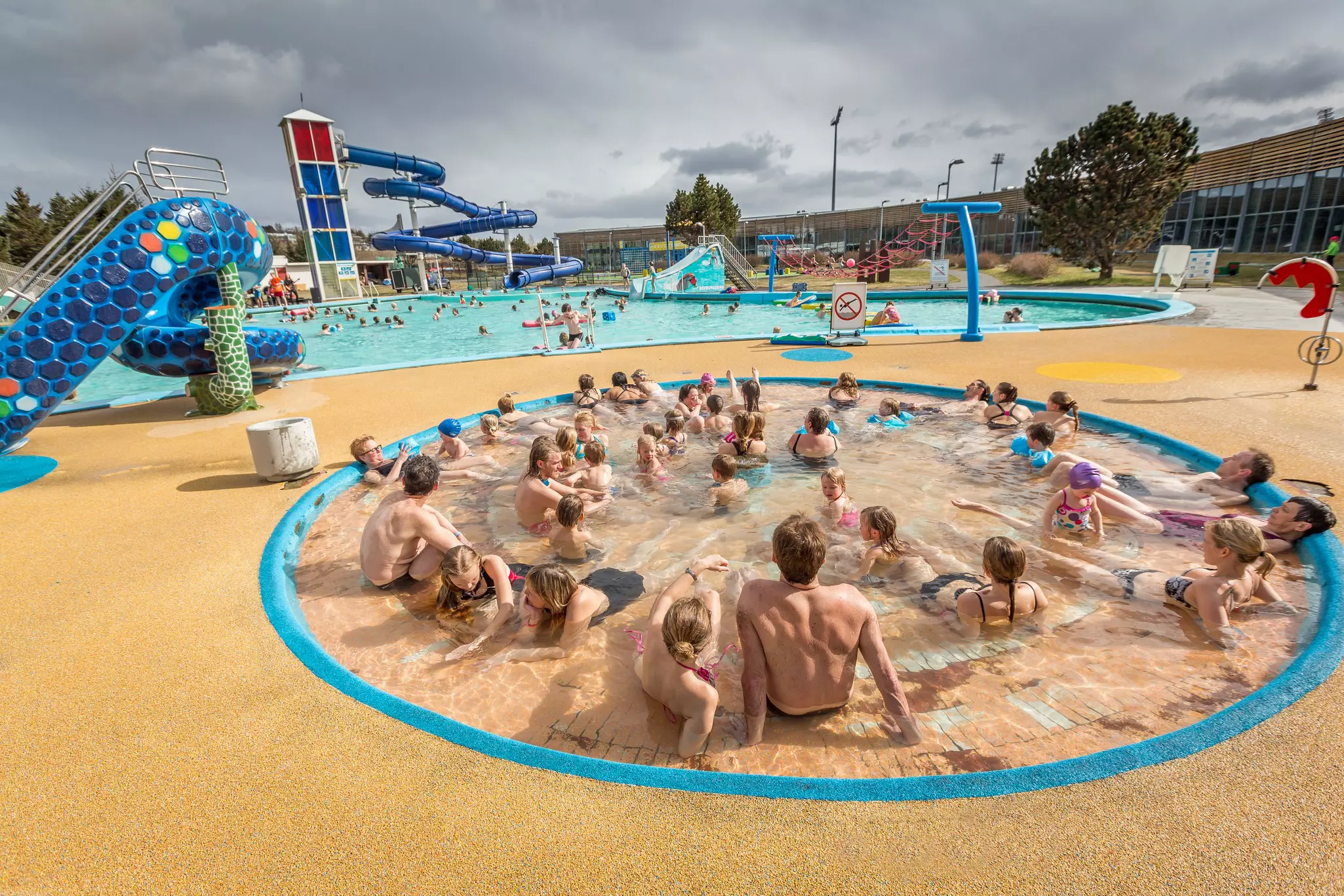 A wide view of children and adults in a shallow outdoor pool at a public pool complex.