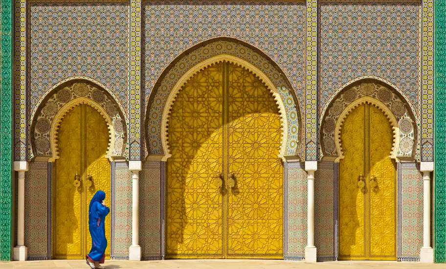 Woman in a blue djellaba walking past the gold doors and mosaic tilework of the Royal Palace gates in Fez, Morocco