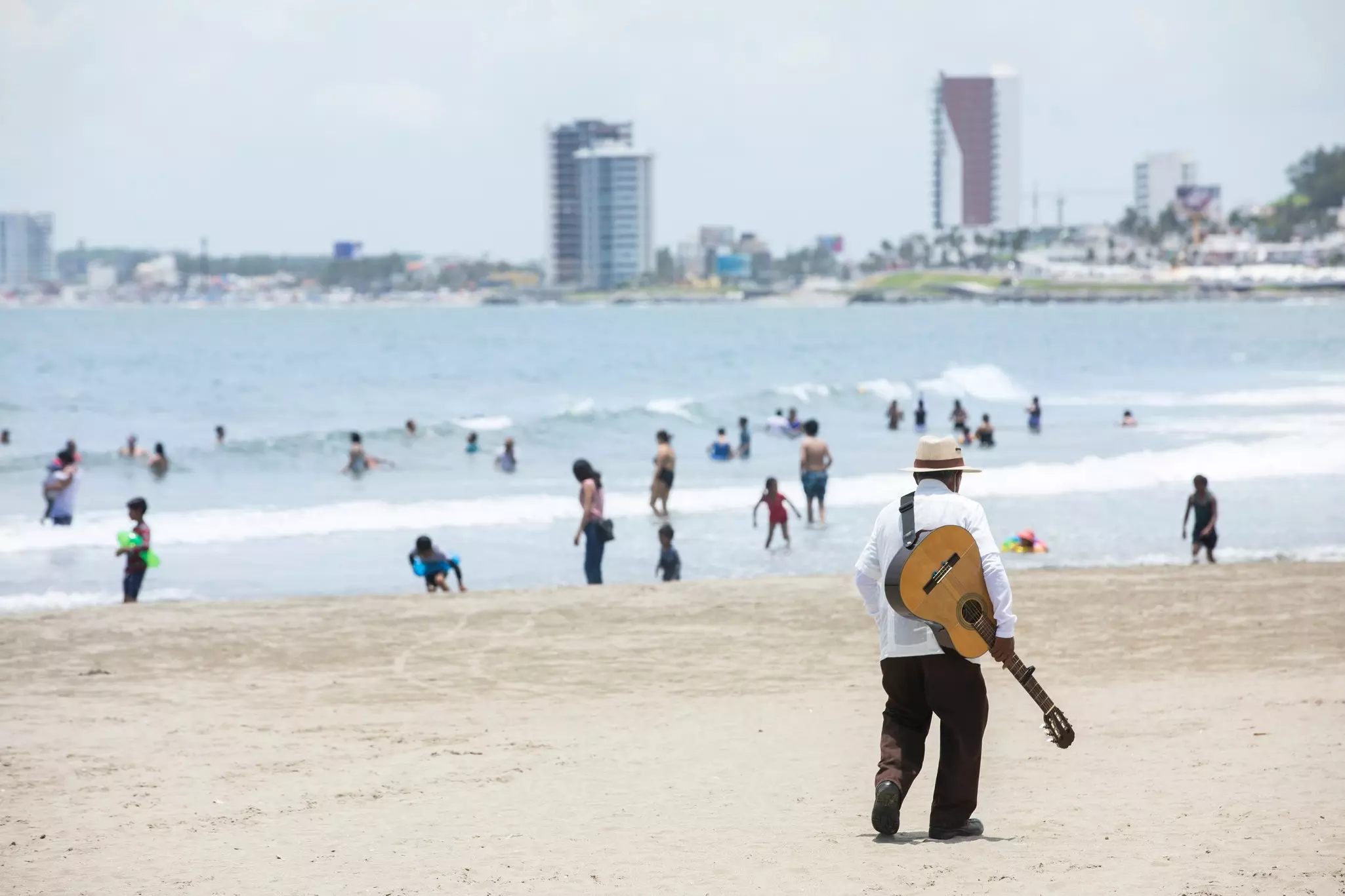 A musician strolls along a beach at midday. Other people are in the ocean and along the shore, and an out-of-focus city skyline can be seen in the distance.