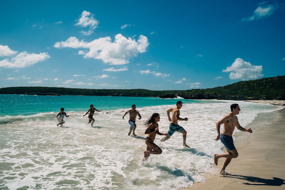 A group of adults and children playfully runs out of the sea with forested hills in the distance on a sunny day.