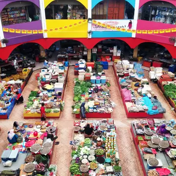 Market stalls piled with vegetables, spices and dried goods on a tiled floor, ringed by painted arches and upper-level shops