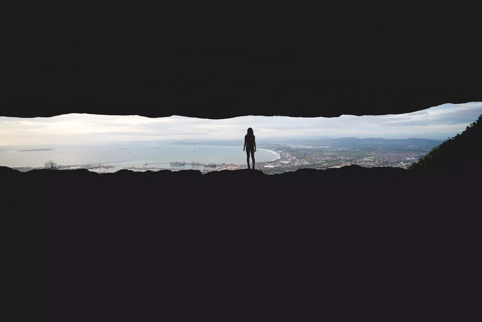 A view through a narrow slit in rocks of a woman's silouette as she looks out over a vast view of a large coastal city. The ocean is in the background.