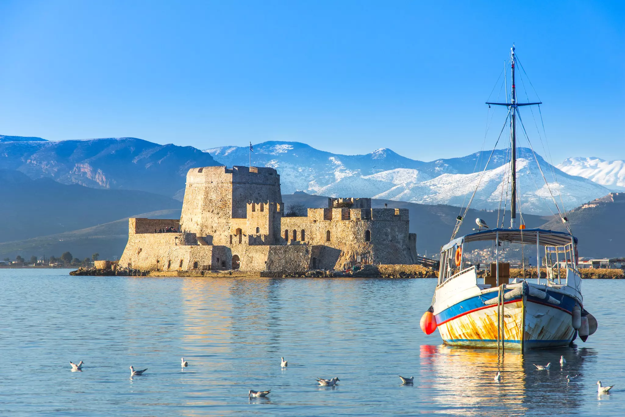 A boat is moored in a harbor near an islet with an imposing stone fortress. Mountains rise on the far shore.