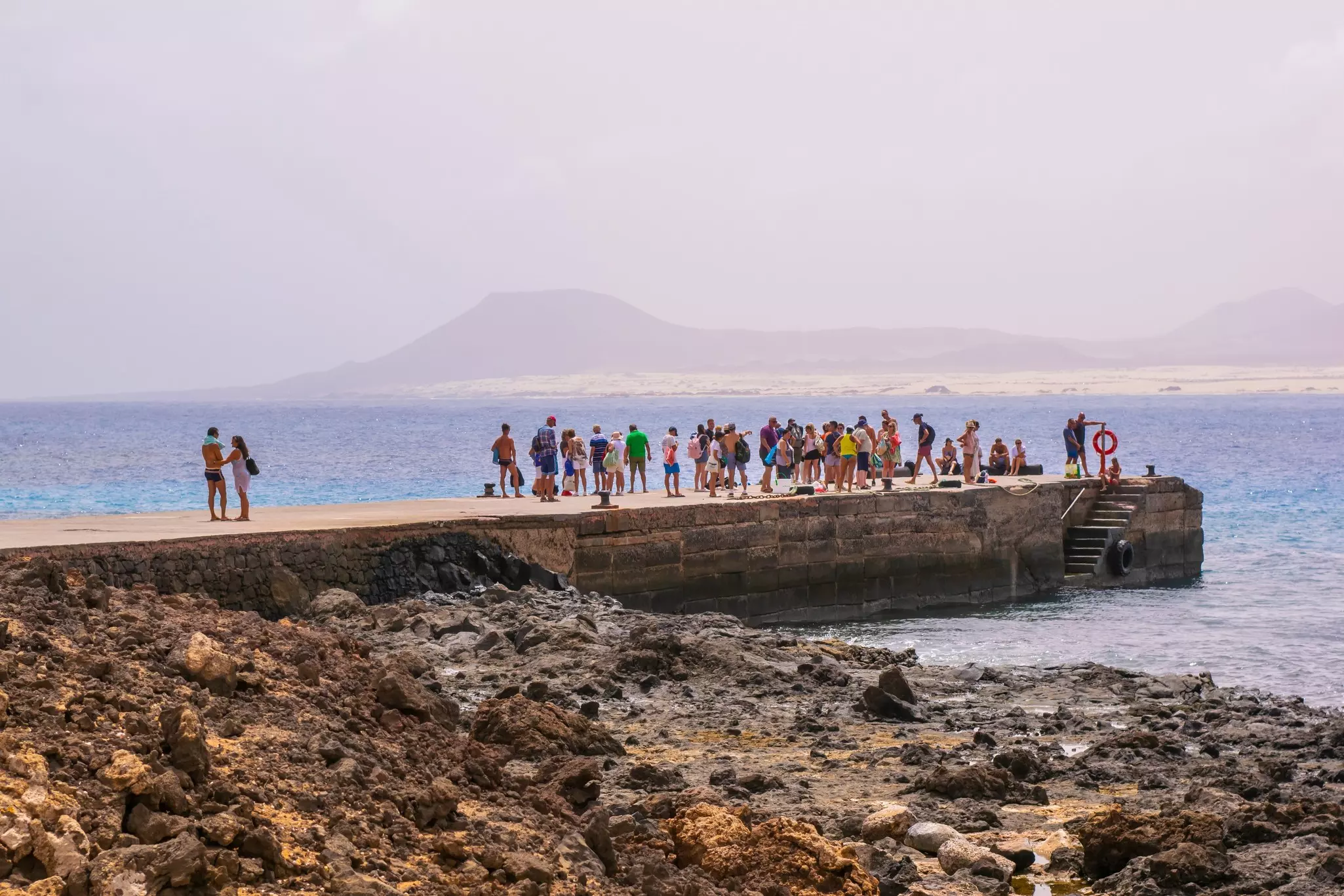 People waiting for a ferry at a small harbour in the sun with beach and volcanic mountains in distance