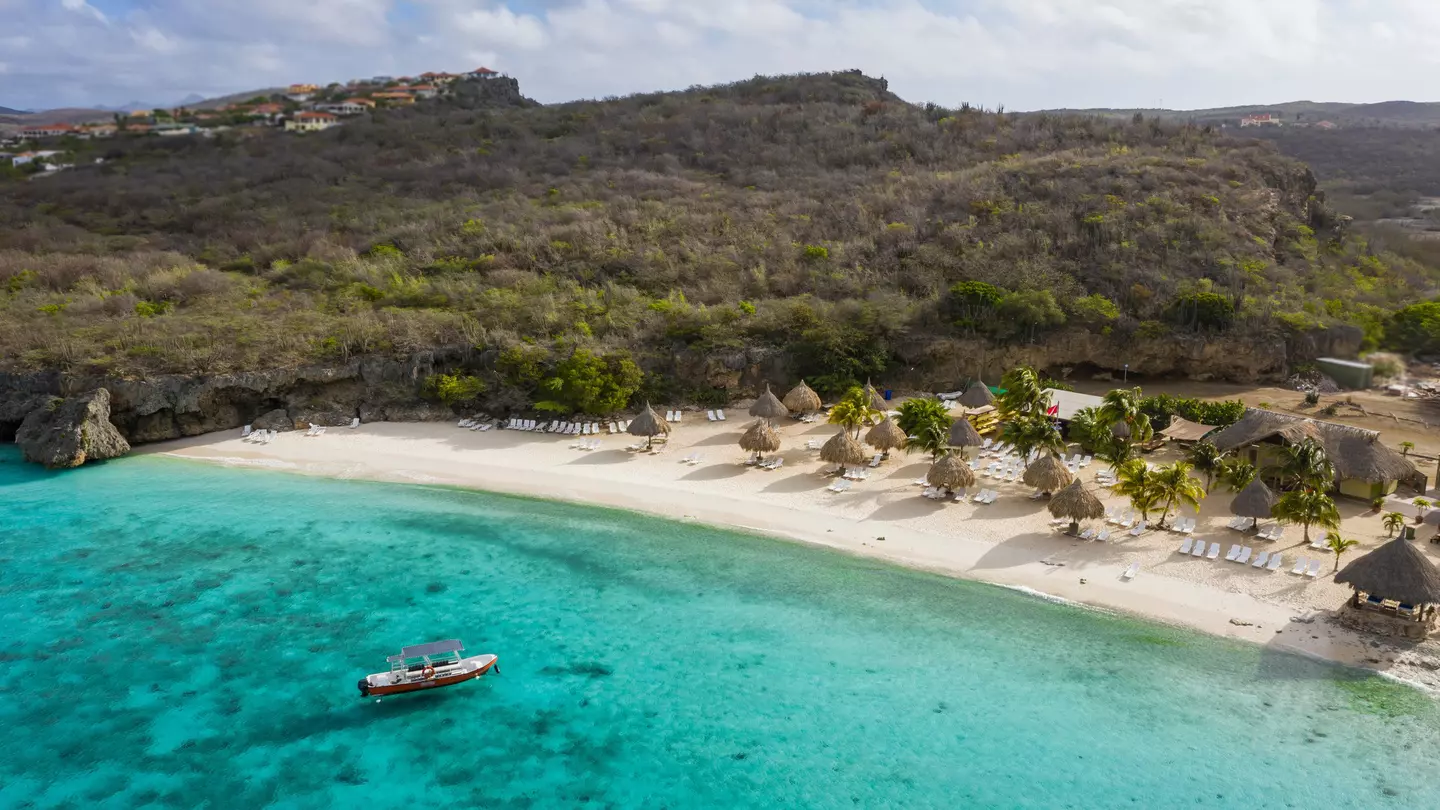 A boat moored in the turquoise ocean near a large beach with sunshades and loungers