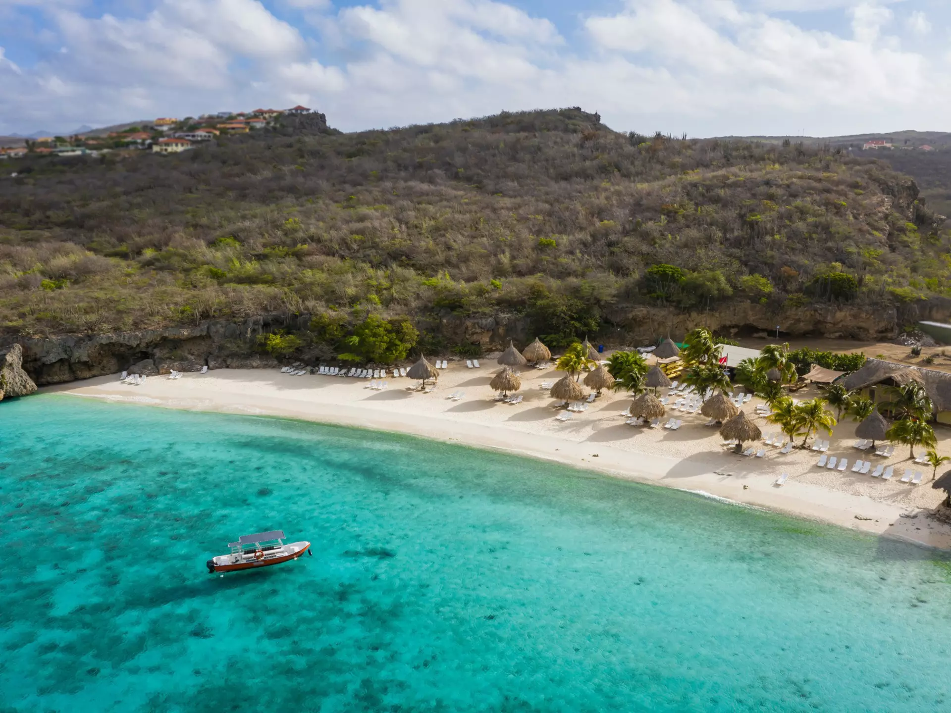 A boat moored in the turquoise ocean near a large beach with sunshades and loungers