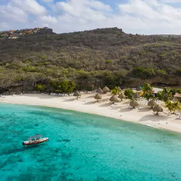 A boat moored in the turquoise ocean near a large beach with sunshades and loungers
