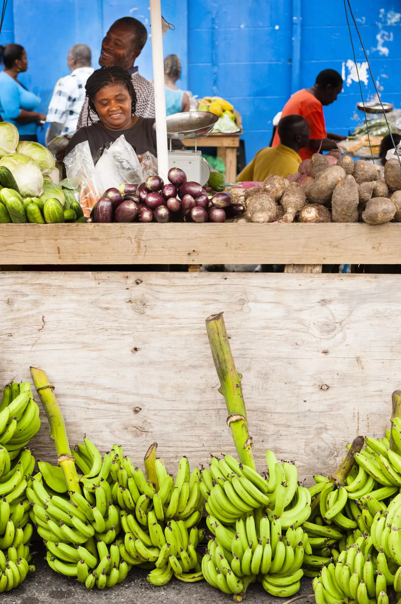 A woman oversees a food stall at a fresh produce market. There are piles of bananas at the foot of the stall.
