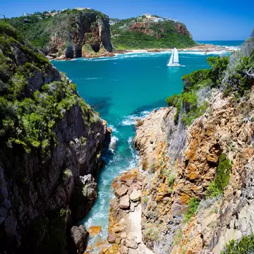 White catamaran on turquoise water between two green headlands, seen down a rocky gully at the Knysna Heads, South Africa.