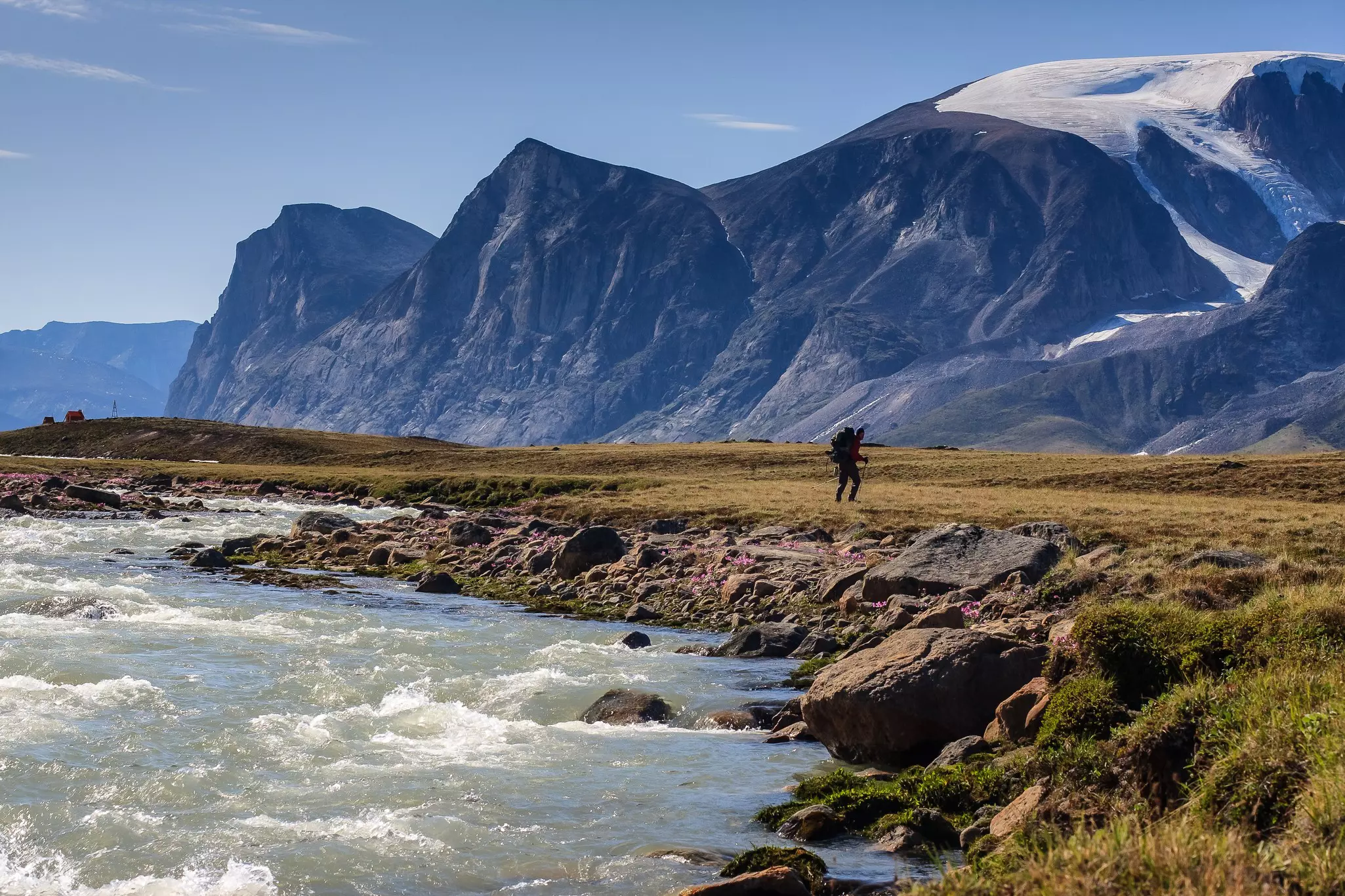 A hiker walks along a rocky shore in a rugged landscape with ice-covered mountains in the distance
