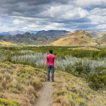 A man in a pink shirt is seen from behind on a hiking trail in a wilderness with meadows and mountains in the distance.