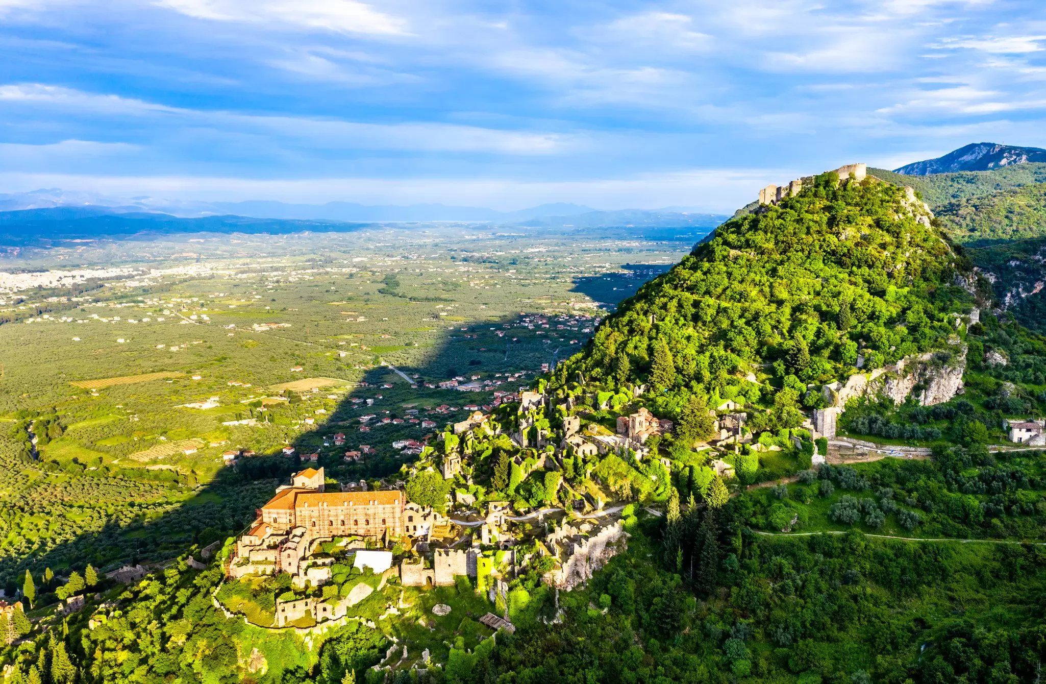 A distant view of the ruins of Mystras in Greece. The ancient stronghold runs up the side of a tall hill, with a castle at the summit.