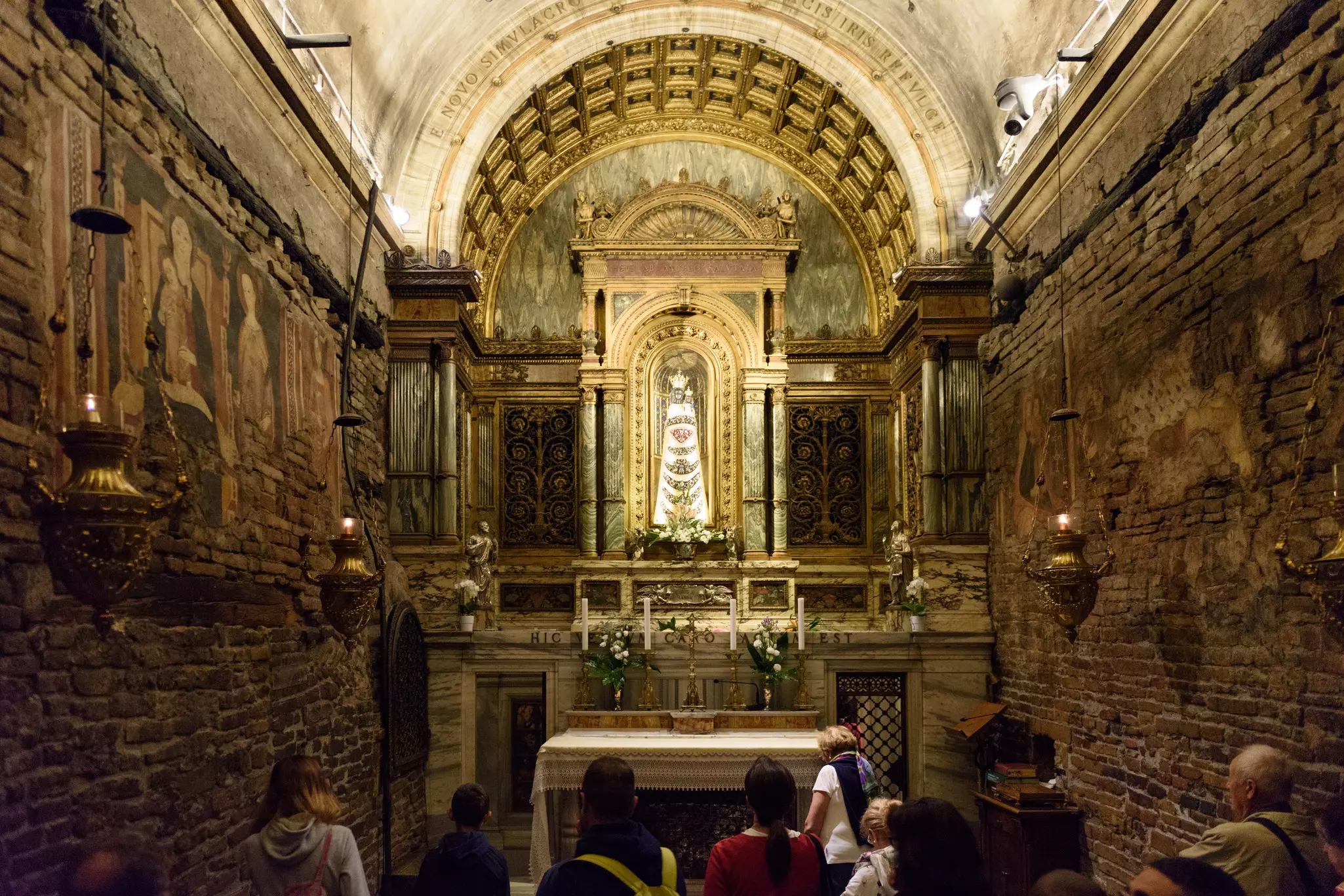 People stand in a narrow room with old brick walls, a vaulted ceiling, and a golden altar at the Basilica Santa Casa. Le Marche, Italy.