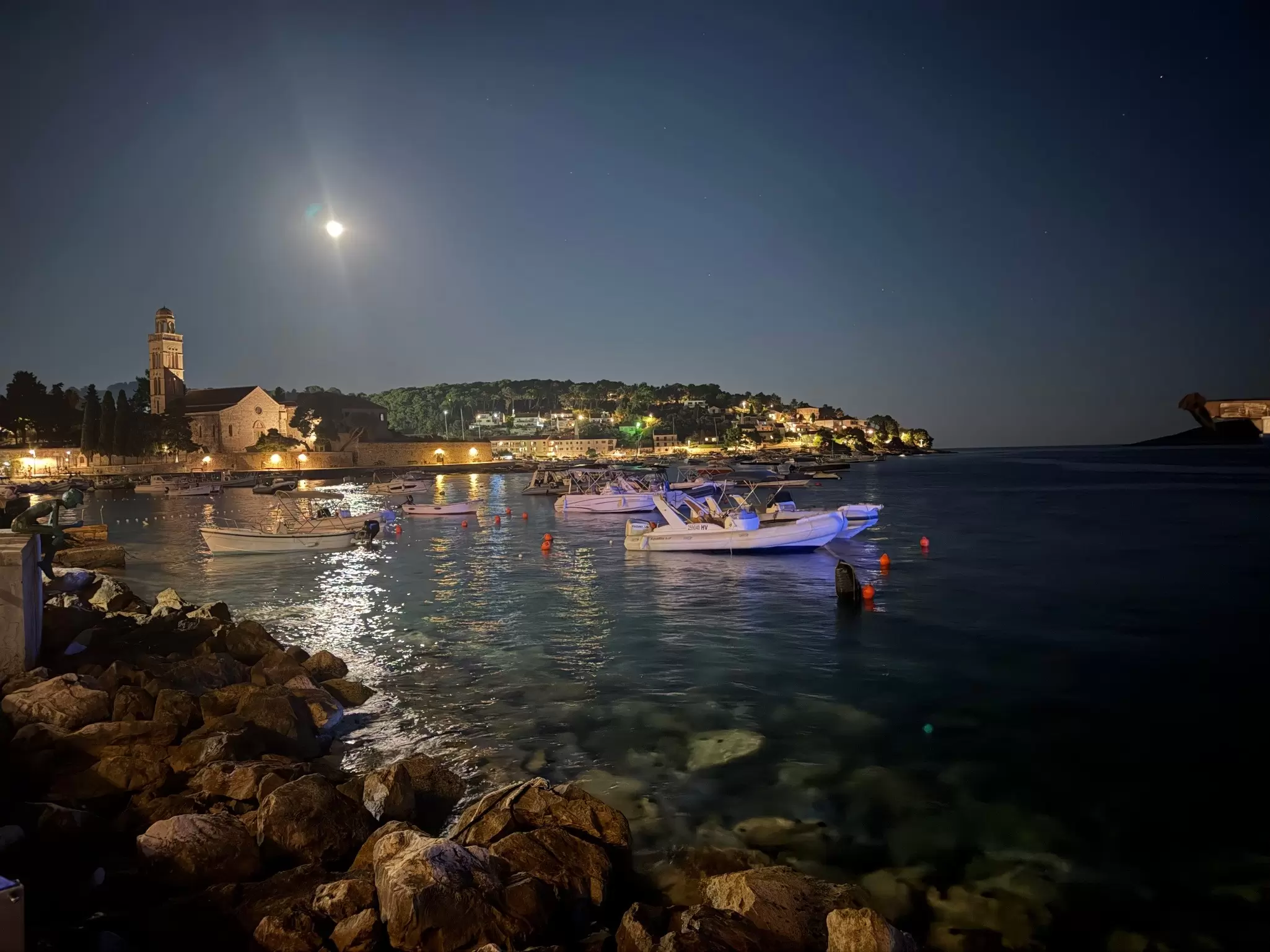 A few boats are moored in the middle of the Adriatic Sea as the moon shines above. Rocks from the shore frame the image.