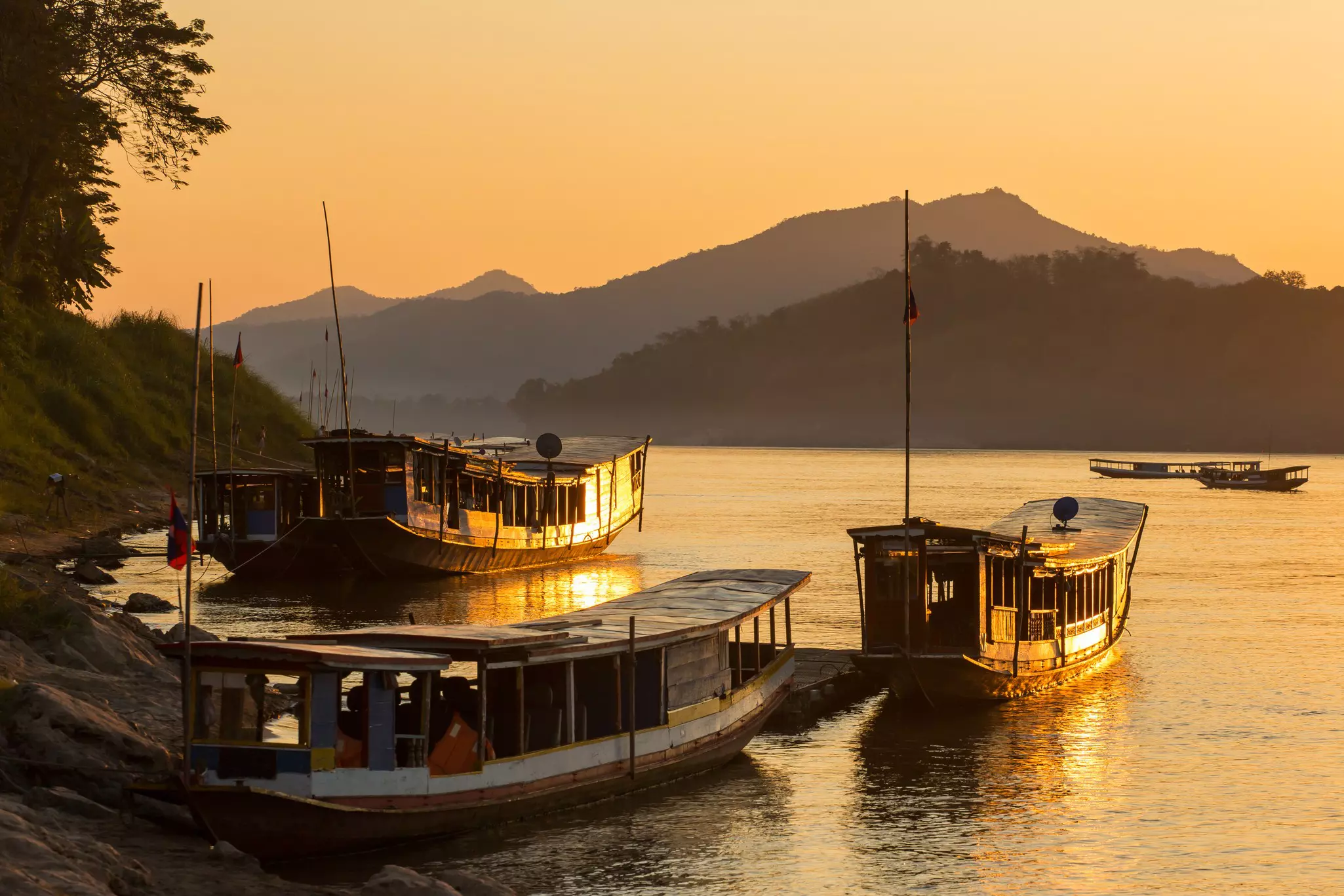 Long, open-sided boats on the shore of a river at sunset. Mountains are in the background.