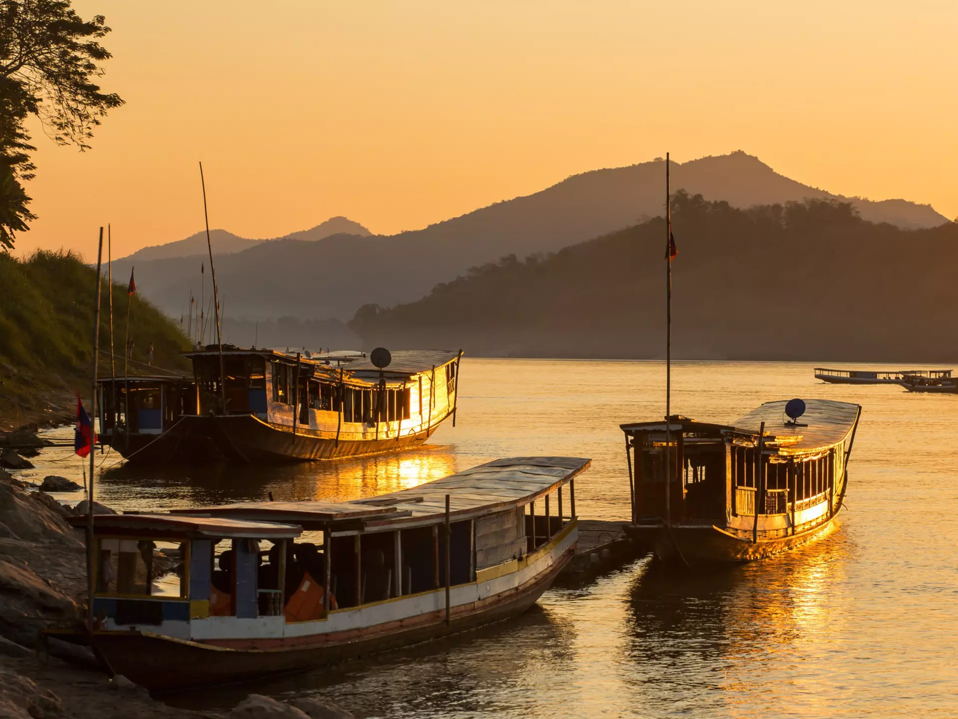 Long, open-sided boats on the shore of a river at sunset. Mountains are in the background.