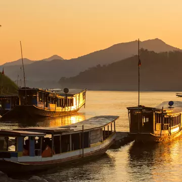 Long, open-sided boats on the shore of a river at sunset. Mountains are in the background.
