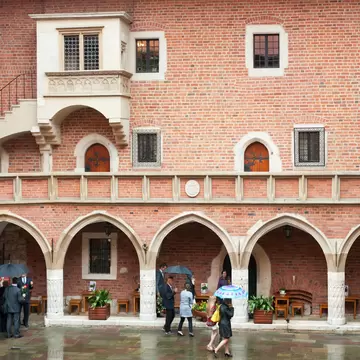 Red-brick courtyard with pointed stone arches, an oriel balcony and visitors holding umbrellas on wet paving.