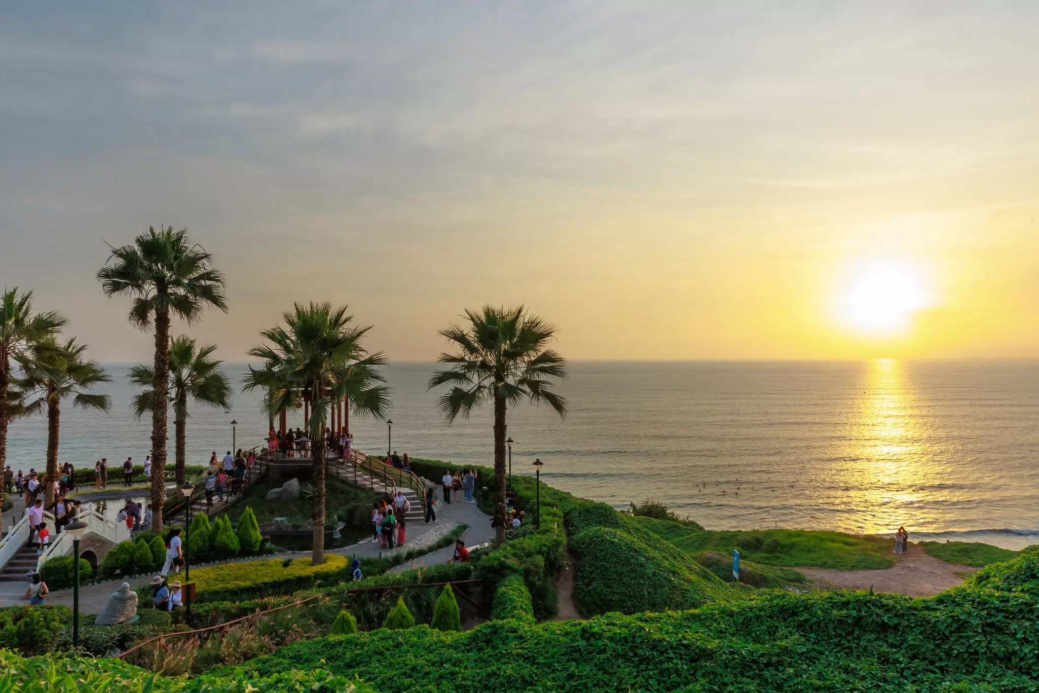 People in a park with palm trees on the waterfront watch the sunset over the ocean.