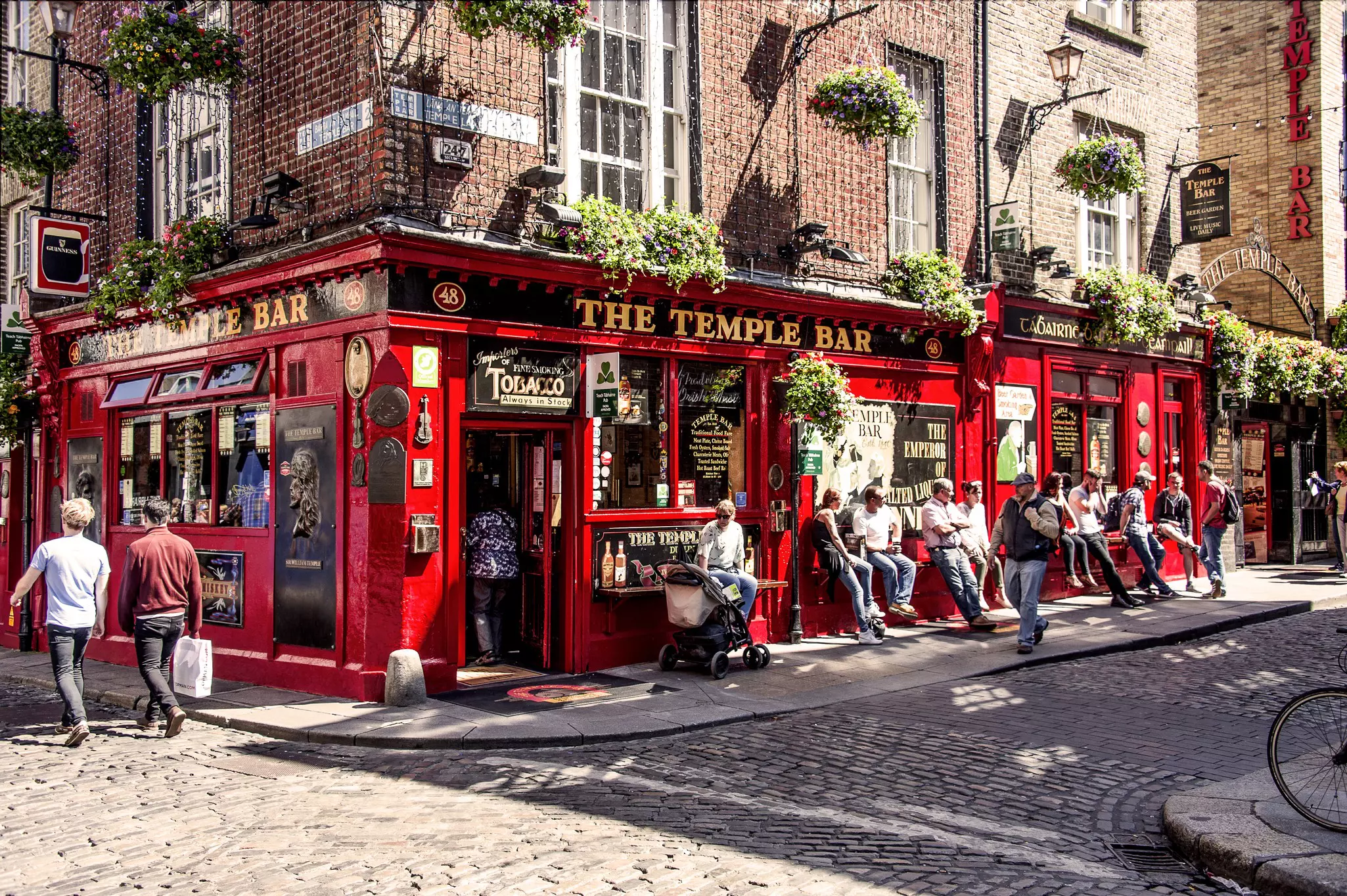 People walking along a cobblestone streeet and sitting outside a corner bar with a bright red facade with hand lettering above the doorway reading "The Temple Bar" on a sunny day.