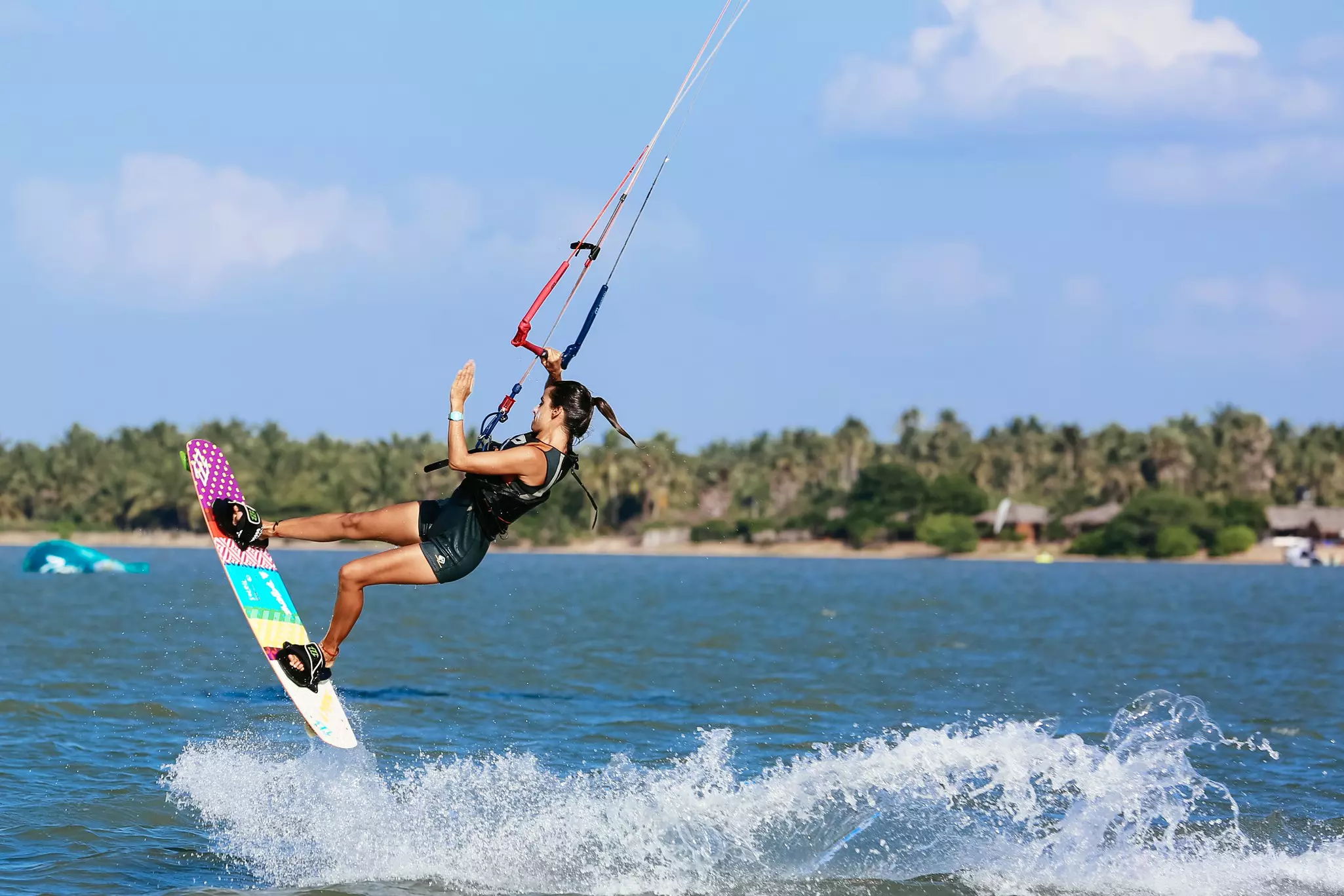 A woman jumps out of the water on a board while kitesurfing off a tropical coast. Whitecaps are seen where her board has cut a wake.